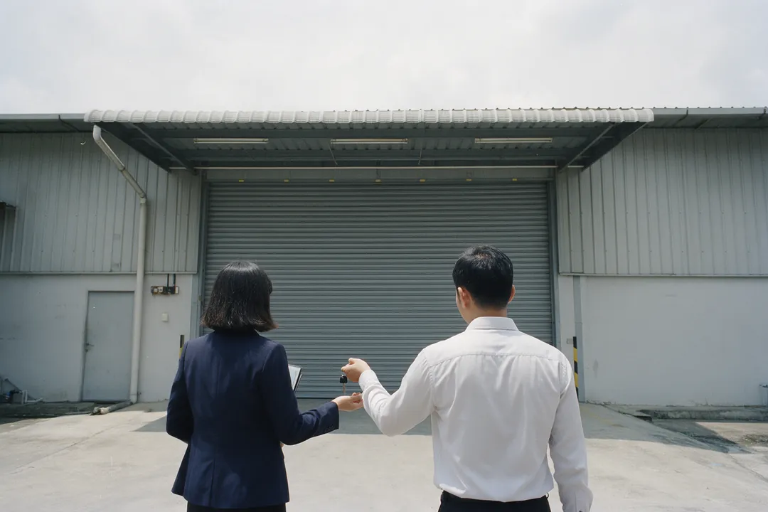 Two people in front of a factory's roller shutter, one handing a set of keys to the other
