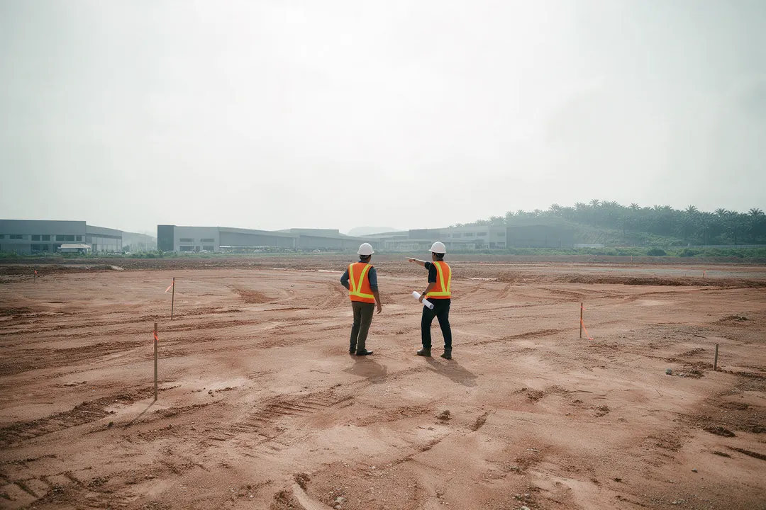 Two people in safety helmets and vests standing on a cleared red-earth industrial plot with survey pegs, new factories in the distance