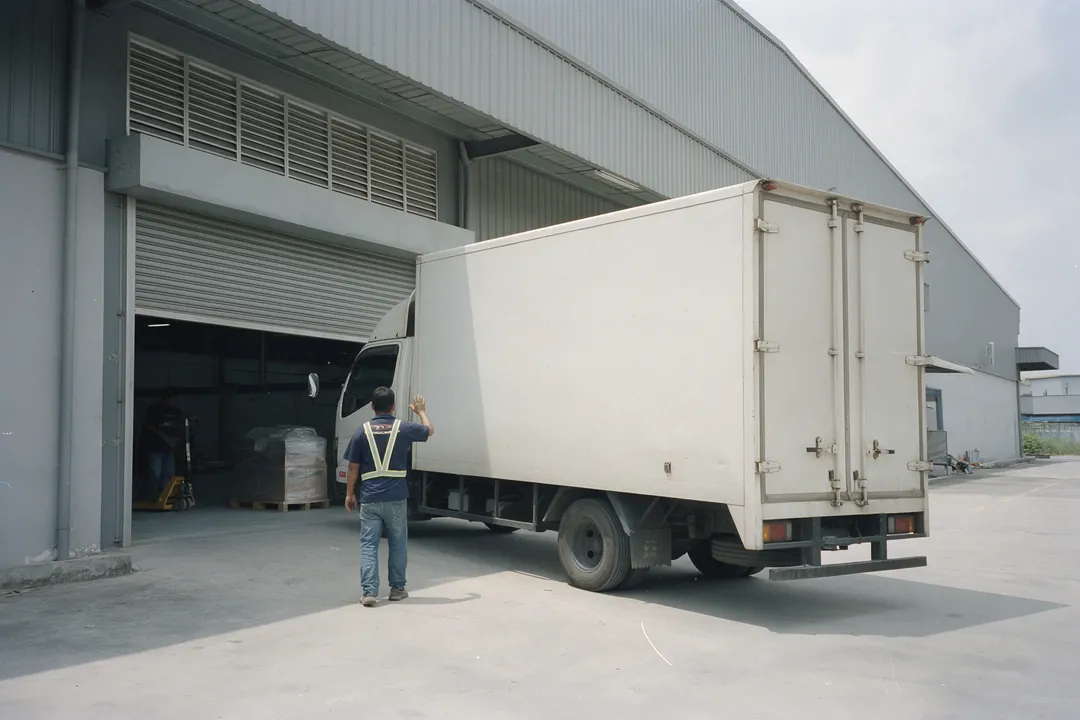 A worker in a safety vest guiding a white lorry towards the open roller shutter of a tall factory, with wrapped pallets inside