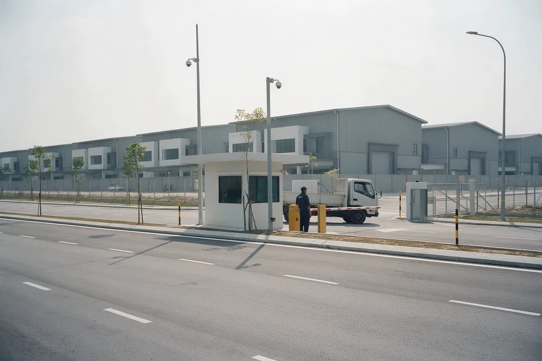 A guard at the barrier of a gated industrial park entrance, with a lorry waiting and new factories behind