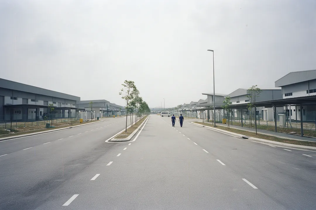 Two workers in navy uniforms walking along a wide new road in a business park, with rows of single-storey factories on both sides
