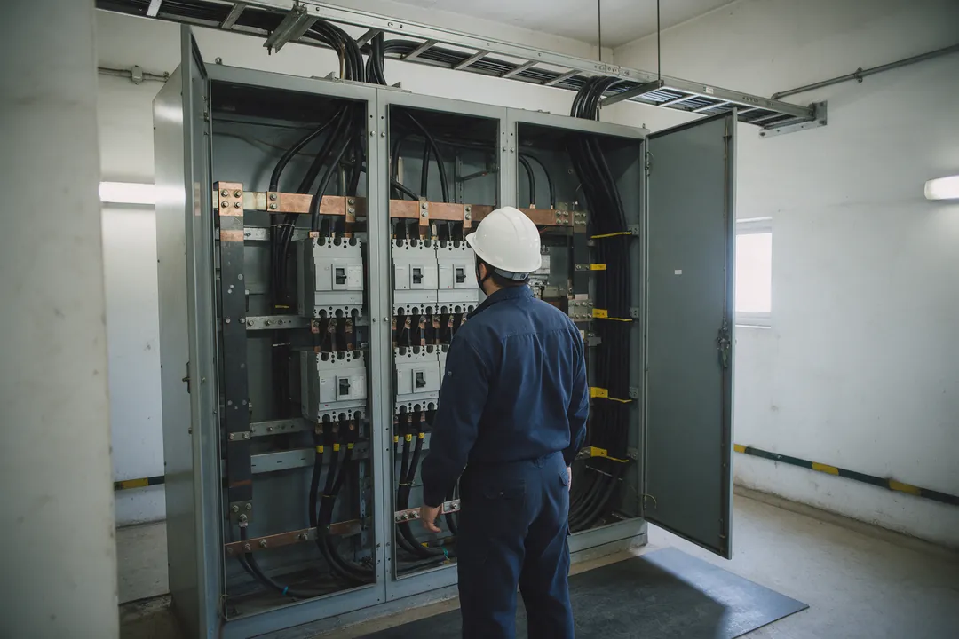An electrician in a navy uniform and safety helmet inspecting an open main switchboard full of circuit breakers and cables