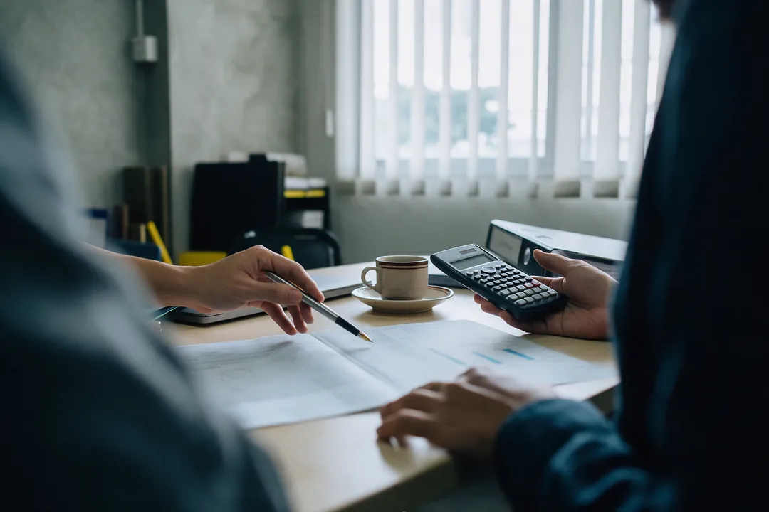 Two people's hands over a printed document on a meeting table, one pointing with a pen, the other holding a calculator